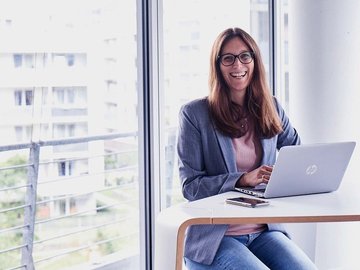 Lächelnde Frau mit langem braunen Haar in einem Büro sitzt vor ihrem Laptop.